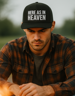 Man wearing a black Christian trucker cap with 'Here As In Heaven' embroidered on the front