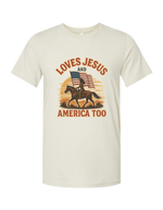 Beige patriotic and Christian t-shirt with graphic of a person on a horse, American flag, and text 'LOVES JESUS AND AMERICA TOO' on a white background.