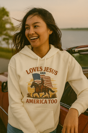 Woman wearing a beige Christian patriotic hoodie with an American flag and text that reads Loves Jesus and America Too.