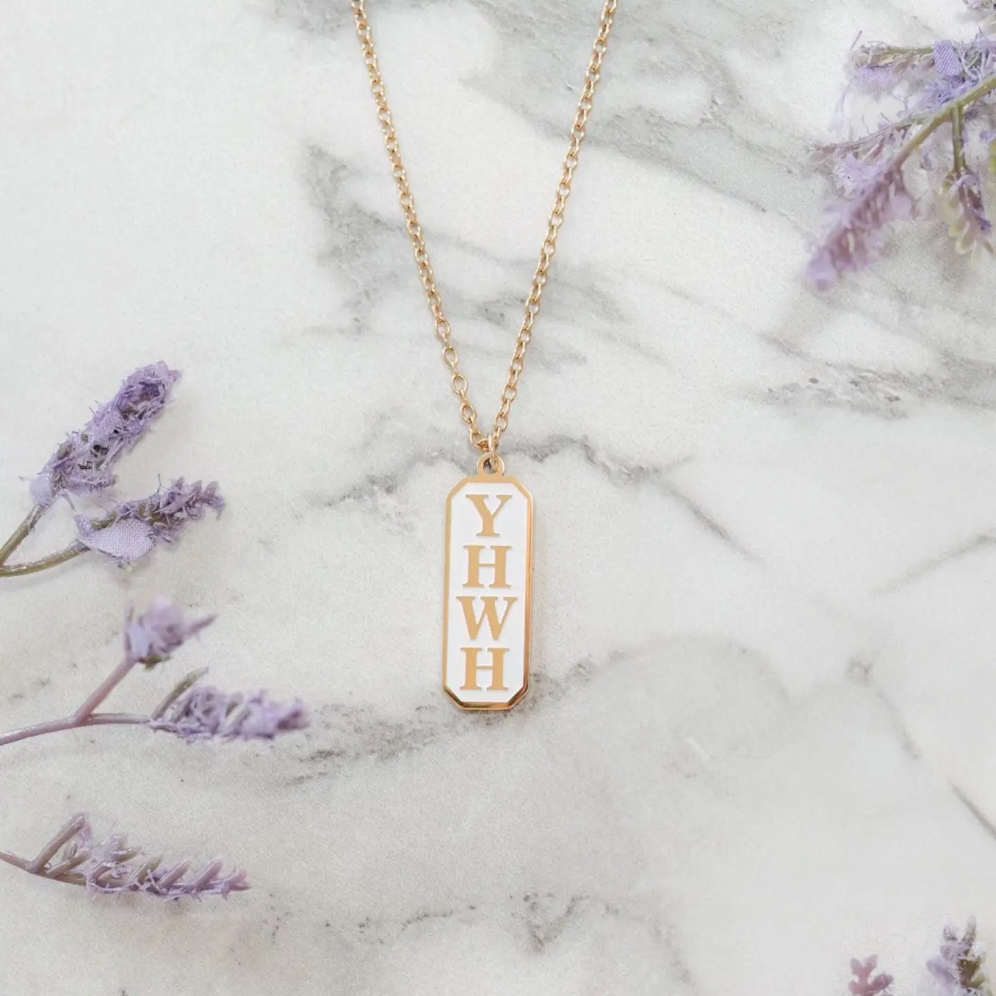 A gold trimmed Christian pendant necklace engraved with 'YHWH' displayed on a marble surface with lavender flowers around it.