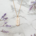 A gold trimmed Christian pendant necklace engraved with 'YHWH' displayed on a marble surface with lavender flowers around it.