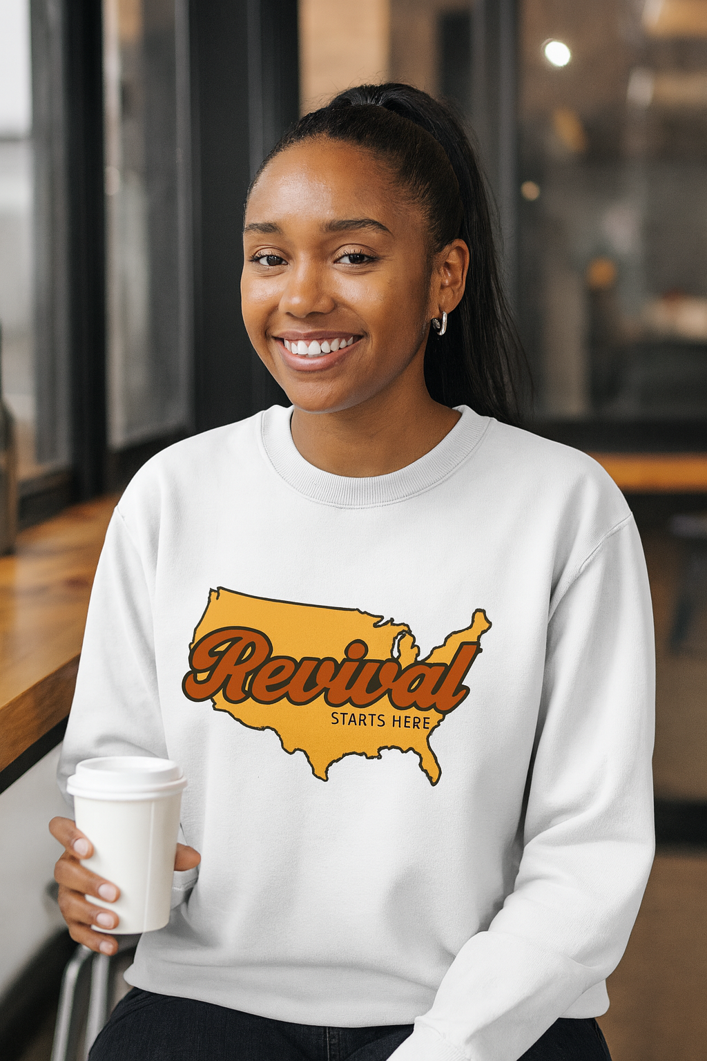 Female wearing a white Christian crewneck sweatshirt featuring Revival Starts Here text overlaying a gold map of the USA. 