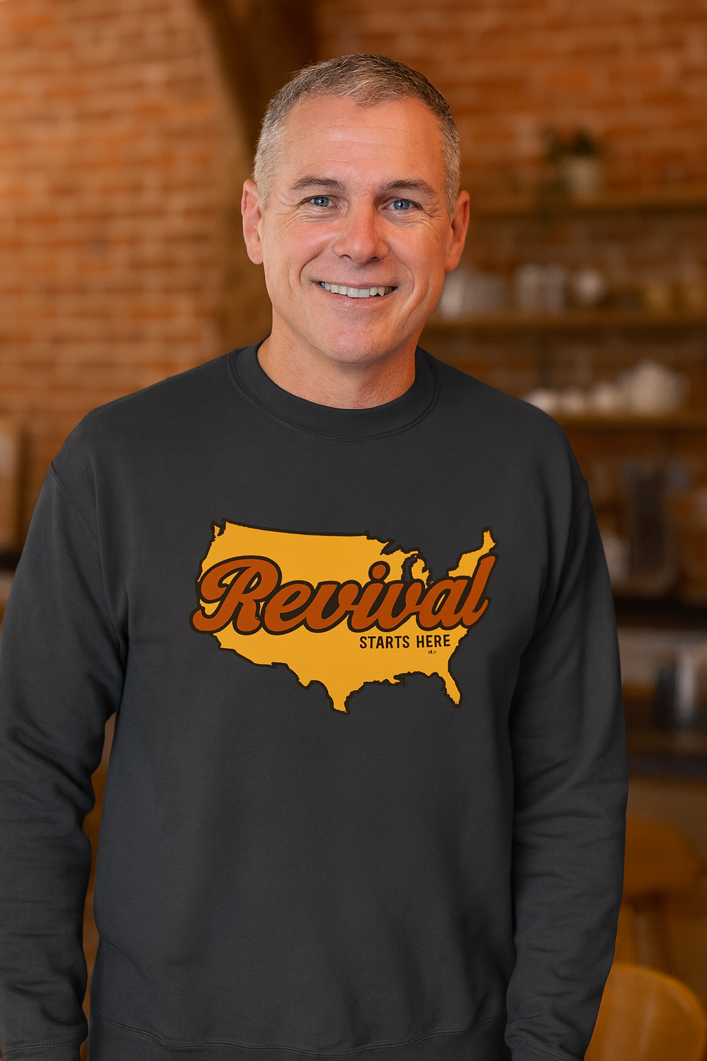 Man wearing a charcoal Christian crewneck sweatshirt featuring Revival Starts Here text overlaying a gold map of the USA. 