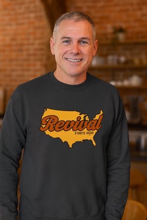 Man wearing a charcoal Christian crewneck sweatshirt featuring Revival Starts Here text overlaying a gold map of the USA. 