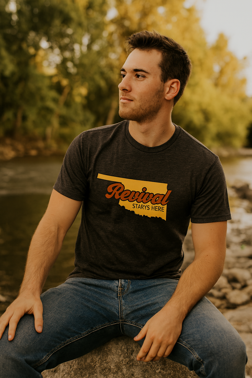 Man wearing a charcoal Christian t-shirt with 'Revival Starts Here' design, sitting by a body of water.