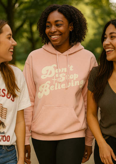 Three women standing outdoors, with one wearing a peach Christian hoodie with text that reads 'Don't Stop Believing.'